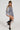 A person poses confidently in the GEMMA Off The Shoulder Voluminous Sequin Mini Dress in Silver, paired with black knee-high boots featuring decorative lace. Their long braided hair cascades down as they stand with one leg crossed in front of the other against a plain background.