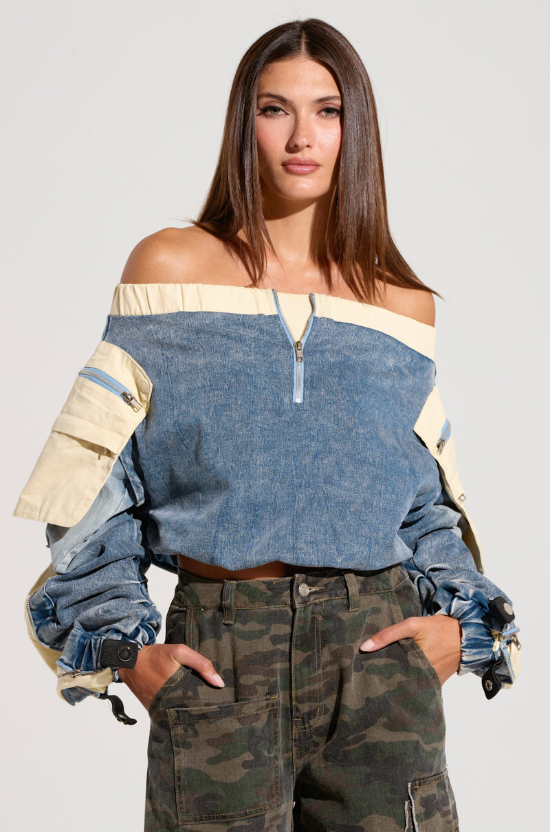 A woman with long brown hair is wearing the CASSANDRA OFF THE SHOULDER DENIM SCRUNCH TOP, featuring a two-toned design with puffy sleeves and metallic snap buttons. She has her hands in the pockets of her camouflage cargo pants, standing against a plain background. 1