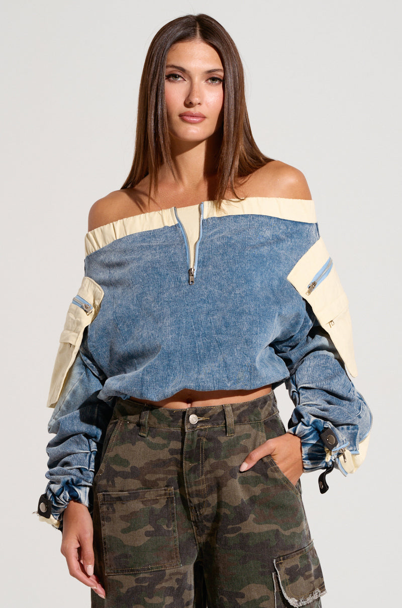 A woman with straight brown hair is wearing the CASSANDRA OFF THE SHOULDER DENIM SCRUNCH TOP in a medium wash, featuring beige accents, a half-zip design, ruched sleeves, metallic snap buttons, and pockets. She pairs it with high-waisted camouflage cargo pants and stands confidently with a neutral expression. 0