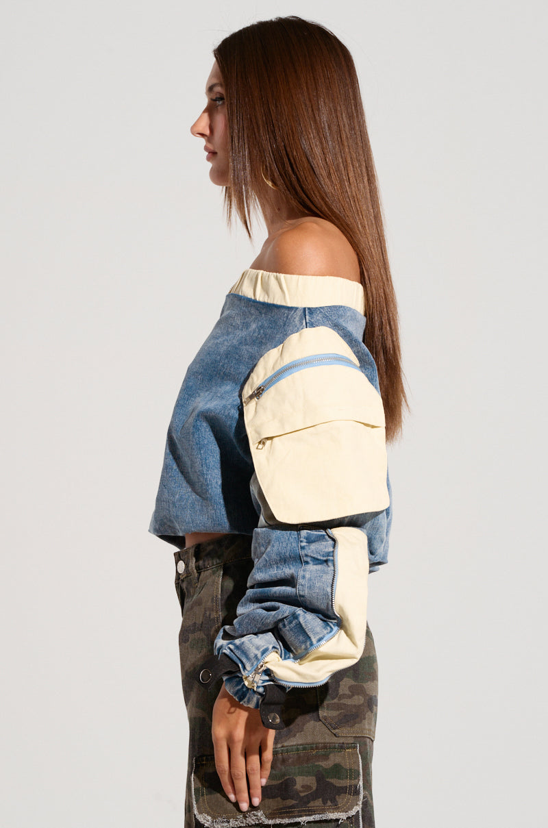 A person with long, straight hair is shown in profile against a plain background. They are wearing the CASSANDRA OFF THE SHOULDER DENIM SCRUNCH TOP, featuring a medium wash with light beige accents and metallic snap buttons. The top has long sleeves and a large pocket. For the lower part, they are dressed in camouflage-patterned pants. 2