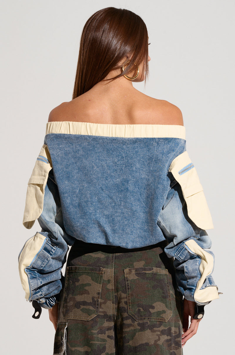 A person with long, straight brown hair is shown from the back. They are wearing the Cassandra Off The Shoulder Denim Scrunch Top with light beige sleeves that have multiple pockets and zippers, paired with camouflage cargo pants. 3
