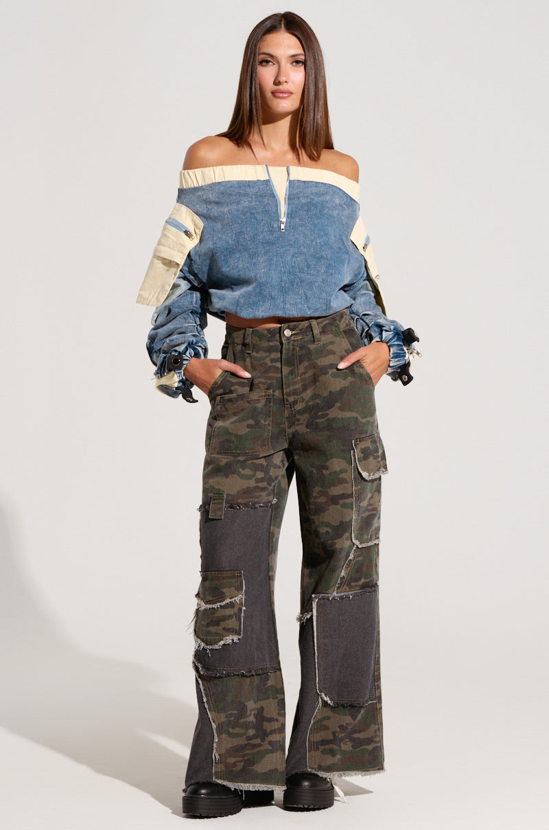 A woman stands confidently with her hands in her pockets, wearing the CASSANDRA OFF THE SHOULDER DENIM SCRUNCH TOP highlighted with cream-colored accents and metallic snap buttons. She pairs it with wide-legged camouflage pants featuring large patches and black platform shoes. 4