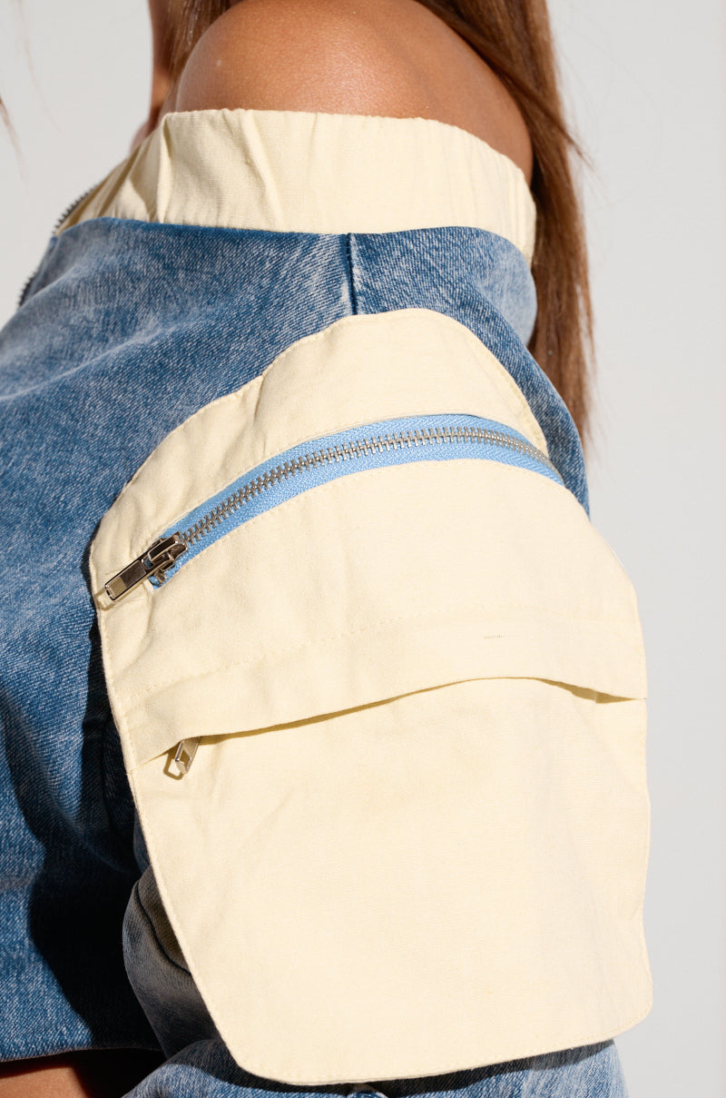 Close-up of a person wearing the Cassandra Off The Shoulder Denim Scrunch Top in medium wash denim, showcasing an attached pale yellow fabric pocket with a light blue zipper. The pocket, positioned on the upper arm, introduces a distinctive and practical element to the garment. 5
