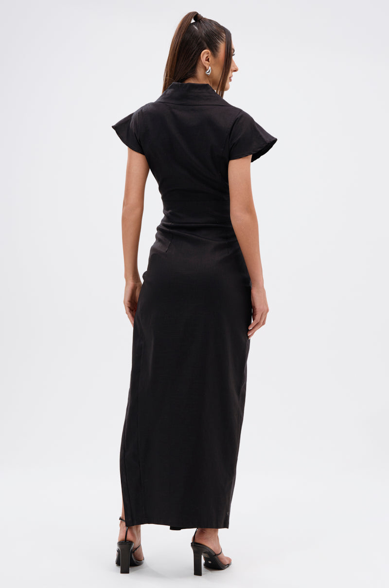 Woman stands with her back to the camera in the CHELSEA BUTTON SIDE MAXI DRESS IN DENIM, paired with black heeled sandals and a ponytail, set against a plain white background.