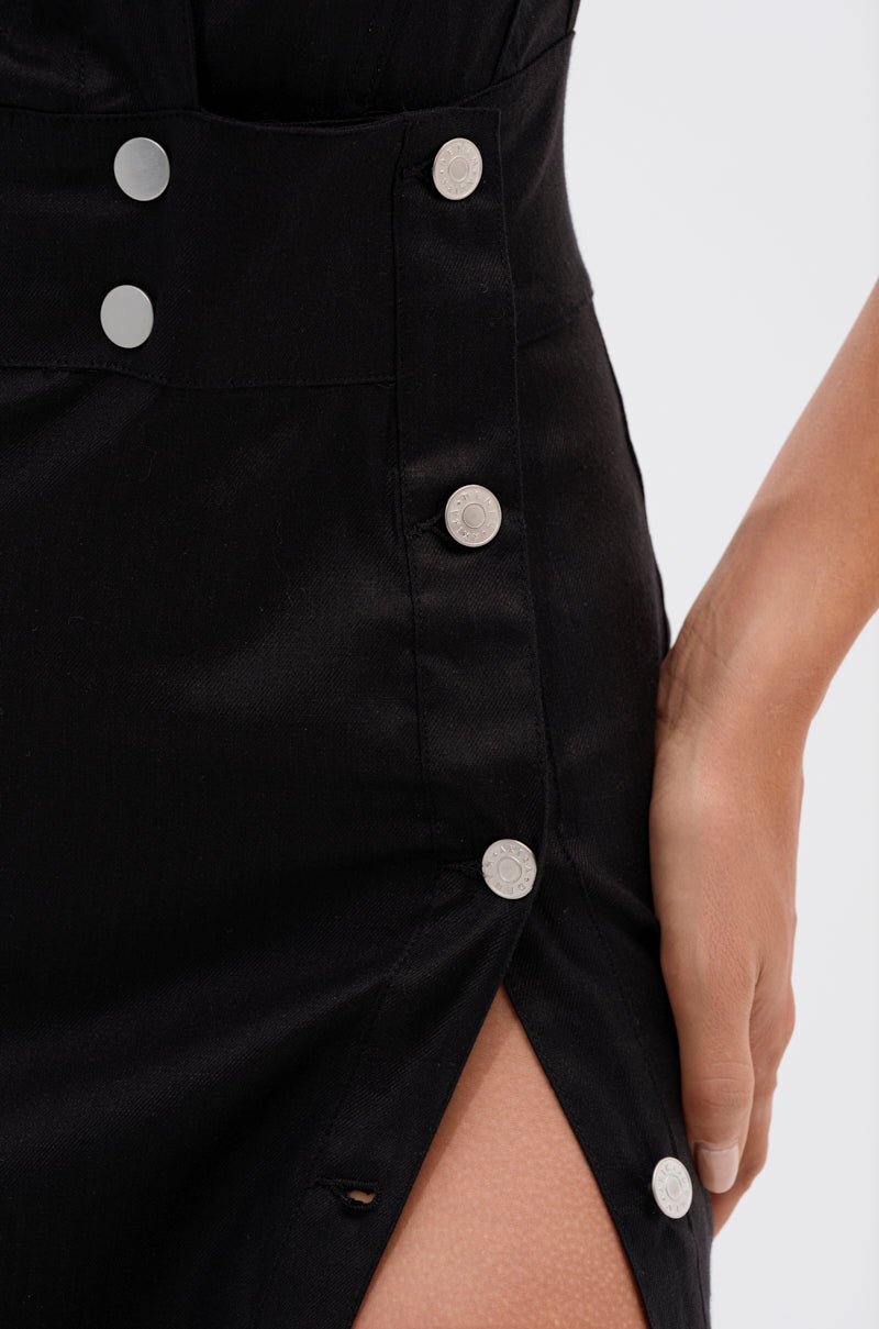 A person wears the CHELSEA BUTTON SIDE MAXI DRESS IN DENIM, featuring silver side buttons and a thigh-high slit. One hand rests near the buttons against a plain, light-colored background.