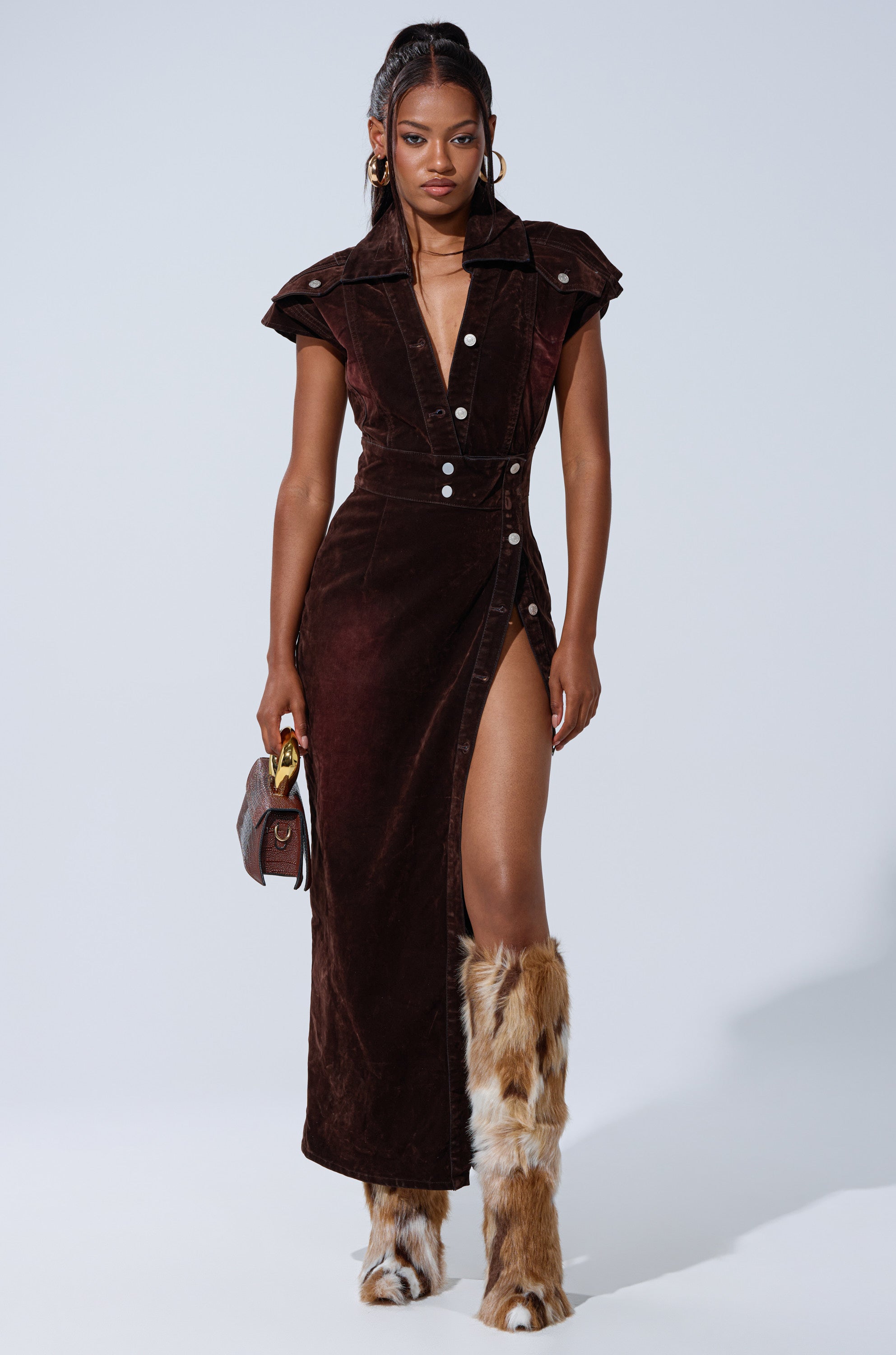 A woman wears the CHELSEA BUTTON DOWN MAXI DRESS IN VELVET DENIM, a dark brown velvet dress with cap sleeves, white buttons, and a deep slit. She pairs it with high furry brown and white boots and carries a small handbag. Her long hair flows in waves.