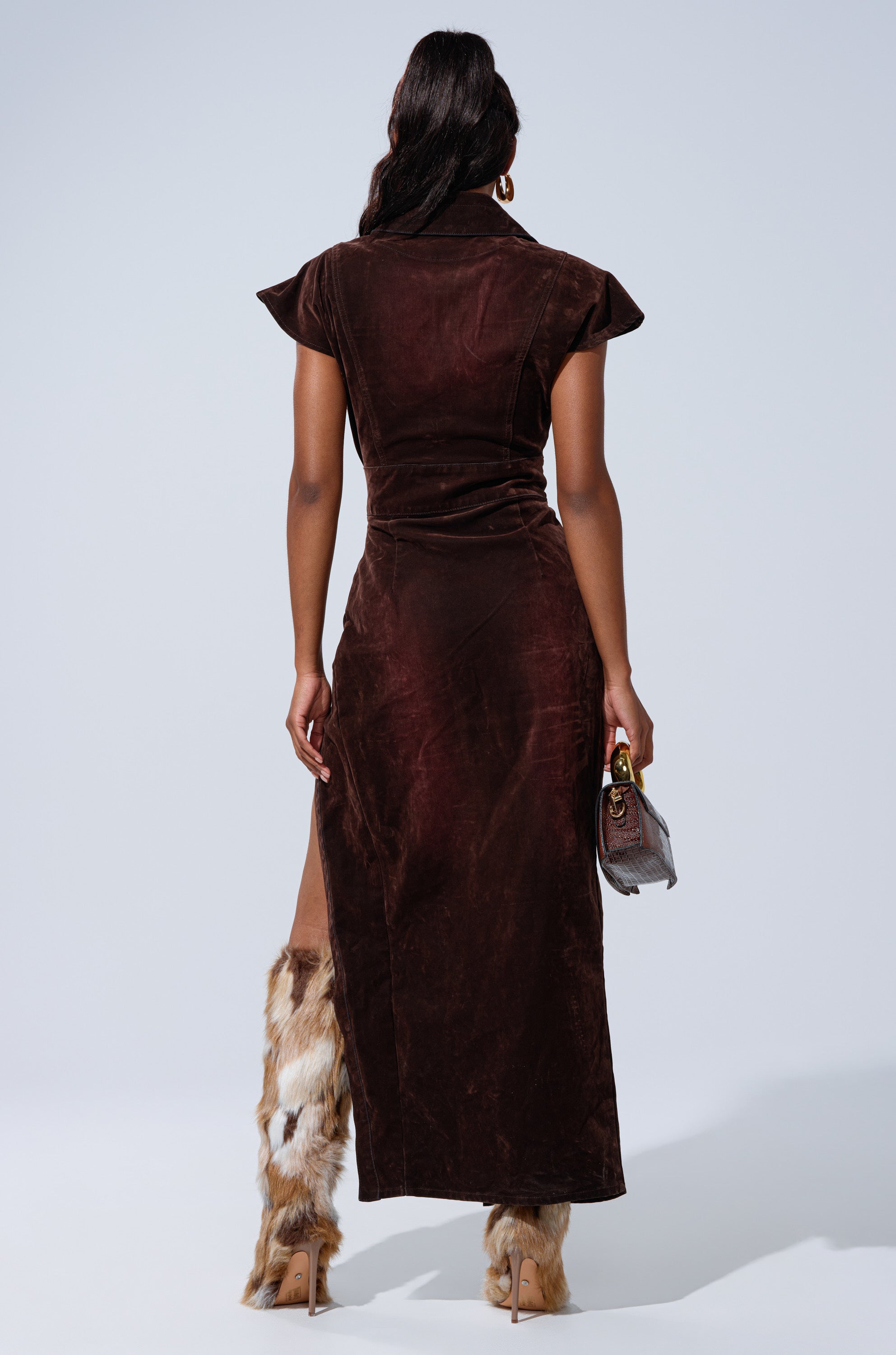 A woman faces away, wearing the CHELSEA BUTTON DOWN MAXI DRESS IN VELVET DENIM, paired with fur-trimmed high-heeled boots and holding a small brown handbag against a plain light background.