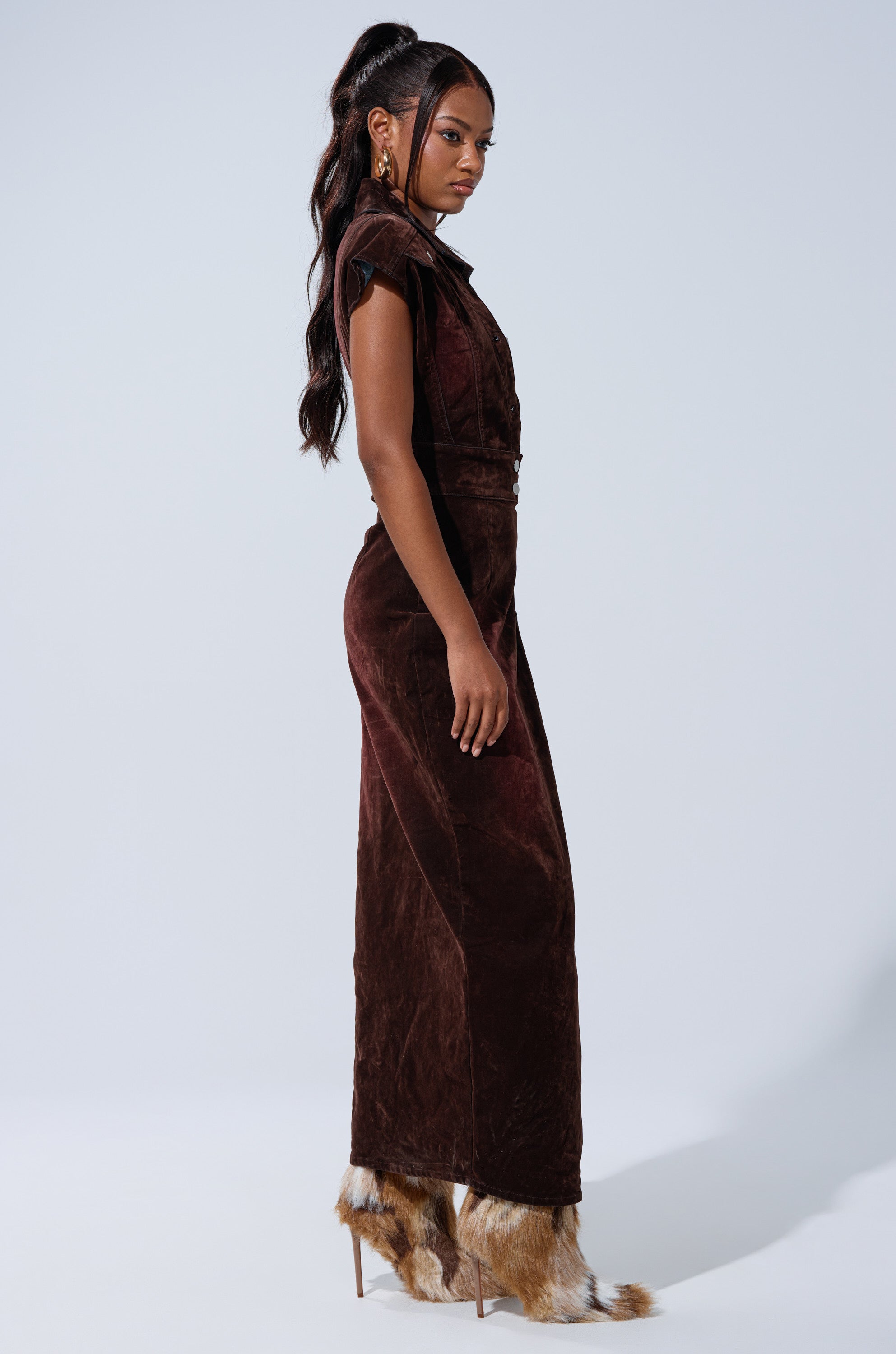 A woman stands sideways in the CHELSEA BUTTON DOWN MAXI DRESS IN VELVET DENIM, paired with furry shoes. Her long wavy hair is tied back, and she wears hoop earrings against a plain, light background.