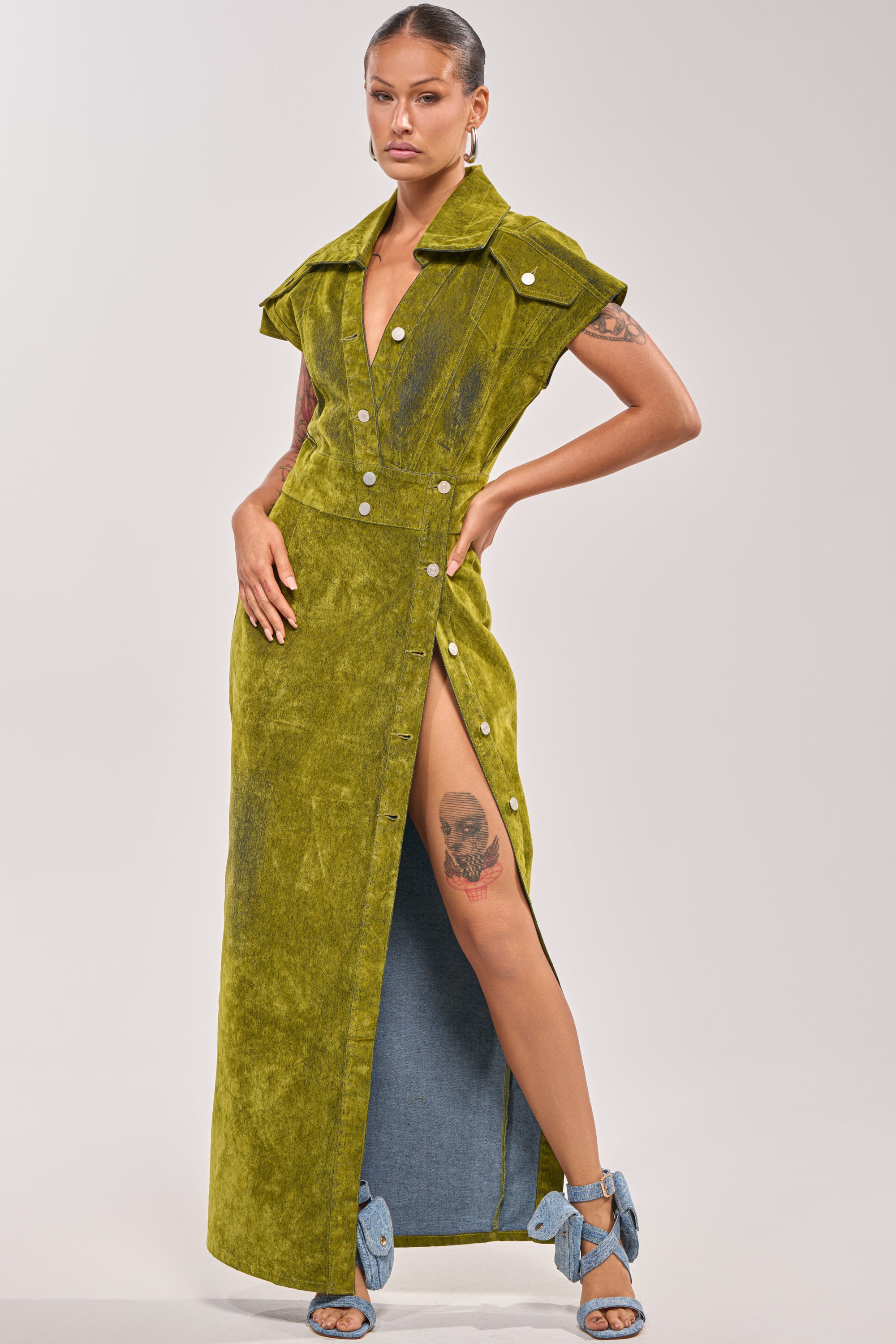 A woman poses in the CHELSEA BUTTON DOWN MAXI DRESS IN CHARTREUSE VELVET DENIM, featuring a sleeveless design, deep V-neck, and thigh-high slit that reveals her skull tattoo. She pairs it with blue heeled sandals with large bows and has her hair pulled back. 0