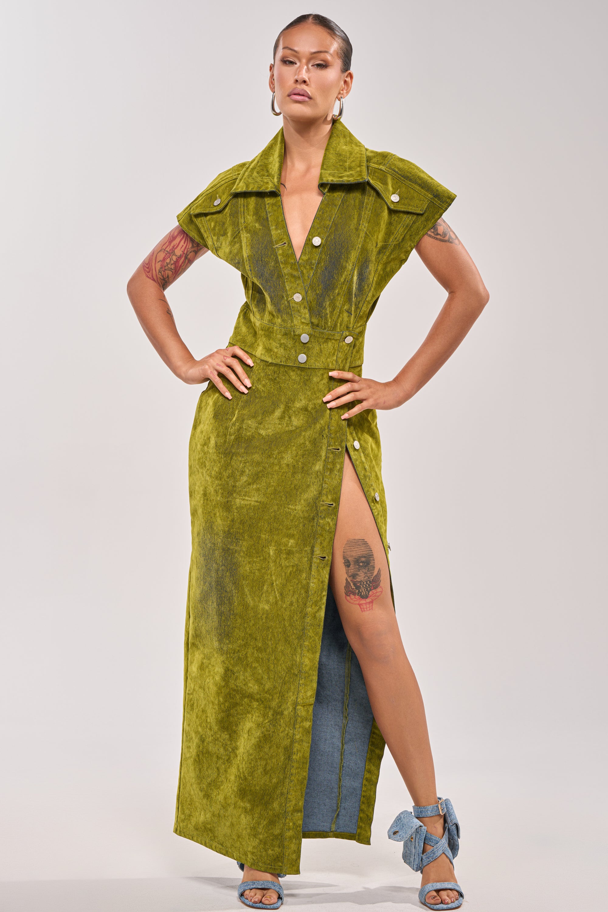 Wearing the CHELSEA BUTTON DOWN MAXI DRESS IN CHARTREUSE VELVET DENIM with a high slit, a person stands confidently, showing thigh and arm tattoos. Their hair is pulled back and they pair the dress with blue-heeled sandals. 1