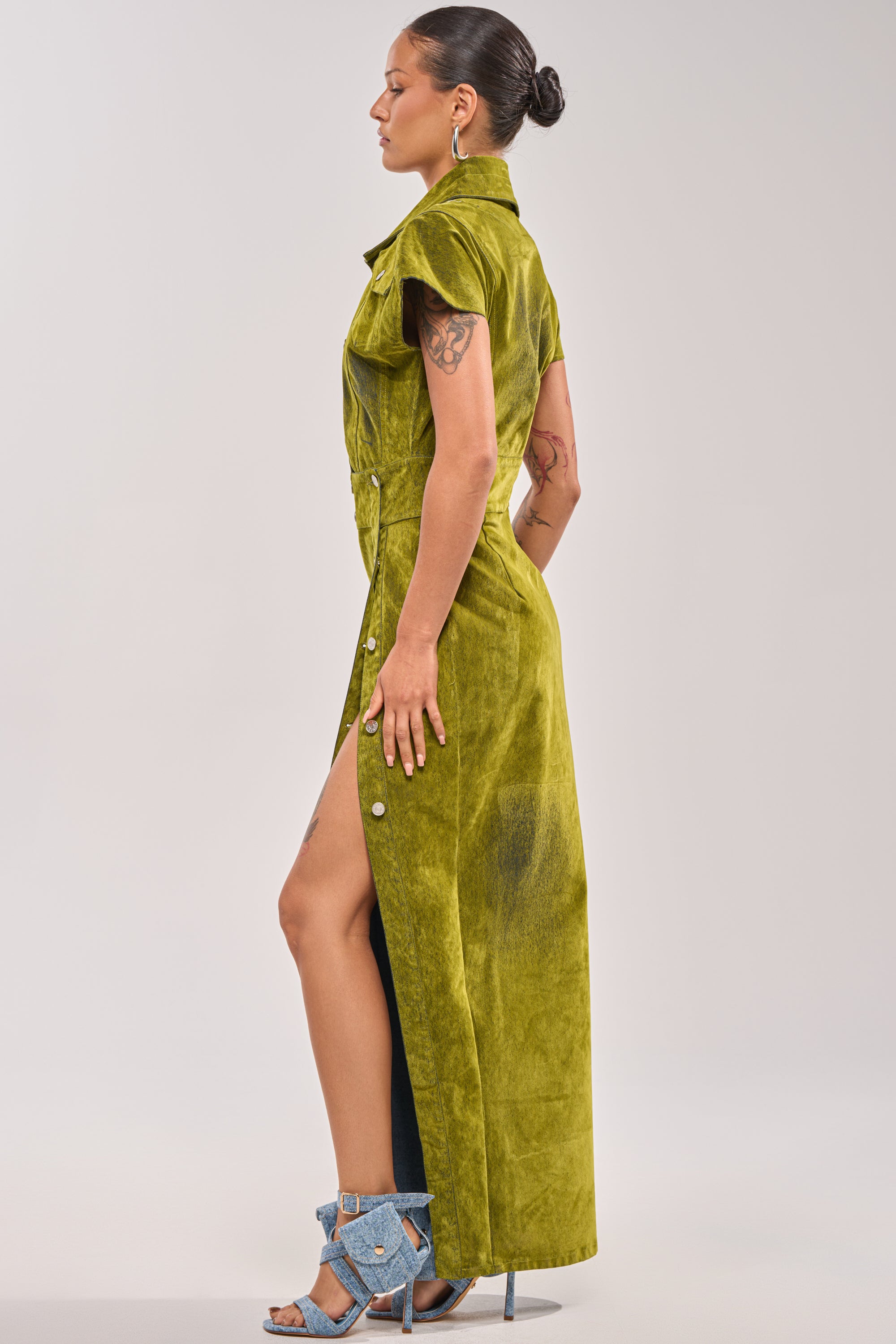 A woman in profile wears the CHELSEA BUTTON DOWN MAXI DRESS IN CHARTREUSE VELVET DENIM with side buttons and high slit, styled with blue heels, arm tattoos, and a bun—perfect for a bold date night look. 2