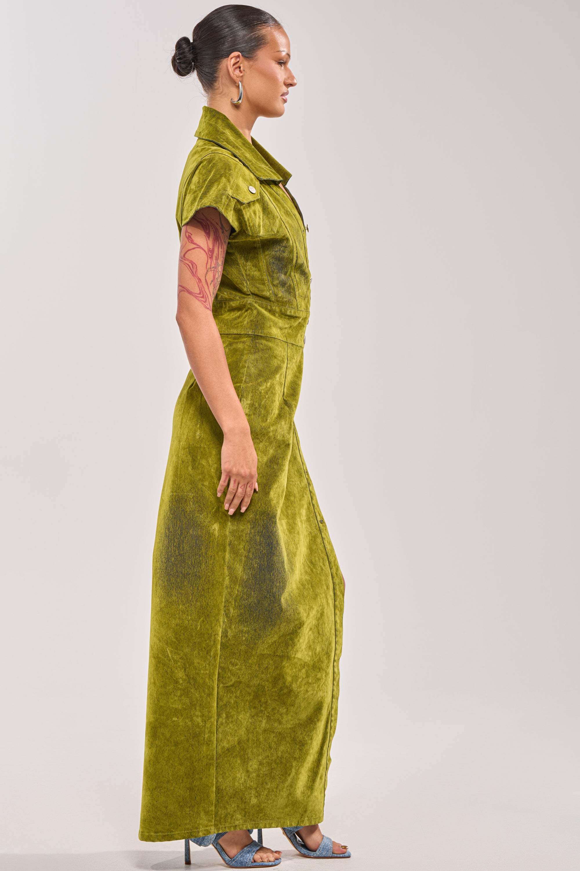 A woman in profile wears the CHELSEA BUTTON DOWN MAXI DRESS IN CHARTREUSE VELVET DENIM; her hair is in a bun, with visible arm tattoos, hoop earrings, and blue high-heeled sandals—an effortlessly chic date night look. 3