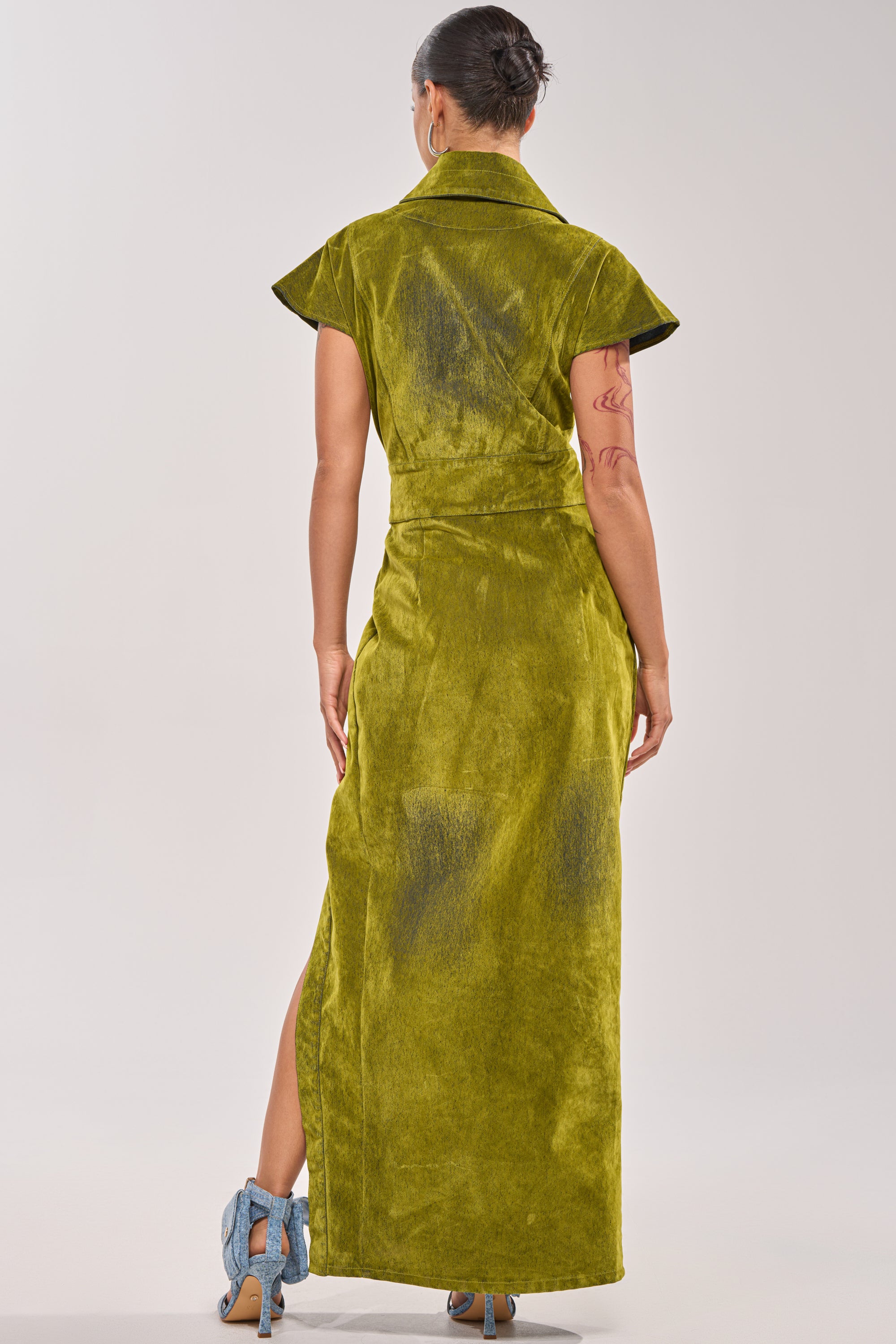 A person faces away, wearing the CHELSEA BUTTON DOWN MAXI DRESS IN CHARTREUSE VELVET DENIM with short sleeves, a collar, and a high slit—perfect for date night—styled with light blue heels and tied-up short hair. 4