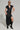 Dressed in a Chelsea Button Side PU Maxi Dress, which highlights its short sleeves and high slit, the individual exudes confidence. The ensemble is elevated by large hoop earrings, sunglasses, and black knee-high boots, standing out beautifully against a simple, minimalistic background.