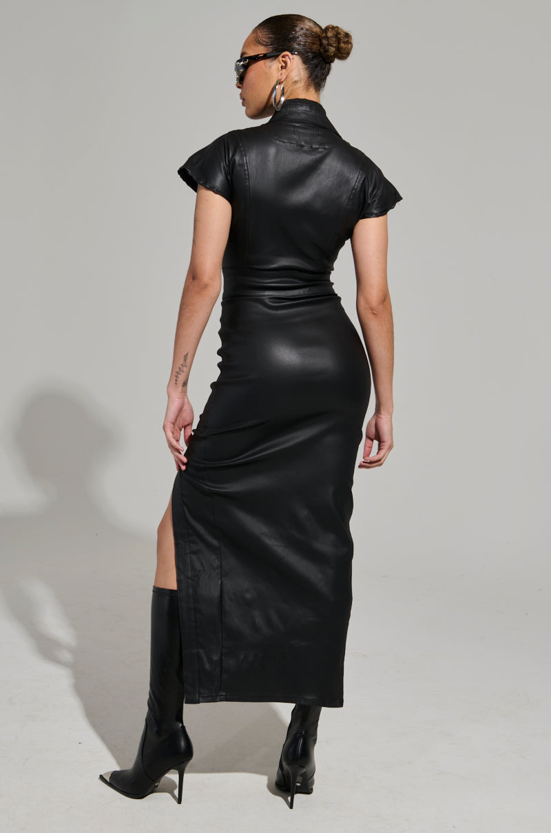 A person wearing sunglasses and sporting a bun is dressed in the CHELSEA BUTTON SIDE PU MAXI DRESS, which accentuates their figure. The faux leather dress features a high slit and complements their knee-high black boots as they stand facing away.