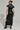 A person wearing sunglasses and sporting a bun is dressed in the CHELSEA BUTTON SIDE PU MAXI DRESS, which accentuates their figure. The faux leather dress features a high slit and complements their knee-high black boots as they stand facing away.