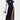 A woman models the CHELSEA BUTTON SIDE DENIM MAXI DRESS, a dark wash maxi with short sleeves, high collar, front buttons, and a bold side slit. She carries a metallic handbag and pairs it with strappy high-heeled sandals.