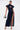 A woman models the CHELSEA BUTTON SIDE DENIM MAXI DRESS, a dark wash maxi with short sleeves, high collar, front buttons, and a bold side slit. She carries a metallic handbag and pairs it with strappy high-heeled sandals.