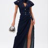 A woman models the CHELSEA BUTTON SIDE DENIM MAXI DRESS, a dark wash maxi with short sleeves, high collar, front buttons, and a bold side slit. She carries a metallic handbag and pairs it with strappy high-heeled sandals.