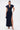 A woman wears the CHELSEA BUTTON SIDE DENIM MAXI DRESS by AKIRA Label, featuring dark denim, cap sleeves, a deep V-neck with high collar, front buttons, and a thigh-high slit. She pairs it with metallic heels and a small handbag, hair tied back.