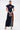 A woman poses against a white background wearing the CHELSEA BUTTON SIDE DENIM MAXI DRESS, featuring a high collar, cap sleeves, front buttons, and a high slit. She accessorizes with a silver purse, gold heels, and hoop earrings.