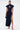 A woman stands against a white background wearing the CHELSEA BUTTON SIDE DENIM MAXI DRESS, featuring a dark wash, high collar, cap sleeves, button side details, and a thigh-high slit. She pairs it with a small handbag and strappy heels, her hair in a sleek ponytail.