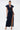 A woman models the CHELSEA BUTTON SIDE DENIM MAXI DRESS featuring cap sleeves, a deep V-neck, large collar, button accents, and a high front slit. She accessorizes with a silver handbag, hoop earrings, and heeled sandals.