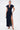A woman poses in the CHELSEA BUTTON SIDE DENIM MAXI DRESS, featuring short sleeves, a collar, and a high slit. She wears gold heels, carries a small handbag, and stands against a white background with her hair pulled back.
