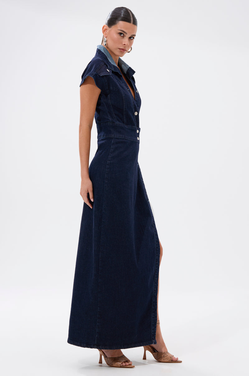 A woman models the CHELSEA BUTTON SIDE DENIM MAXI DRESS in dark blue with a collar, front buttons, short sleeves, a high slit, and tan heeled sandals against a plain white background.