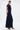 A woman models the CHELSEA BUTTON SIDE DENIM MAXI DRESS in dark blue with a collar, front buttons, short sleeves, a high slit, and tan heeled sandals against a plain white background.