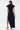 A woman stands facing away, wearing the CHELSEA BUTTON SIDE DENIM MAXI DRESS in dark blue with a high side slit. She carries a small white handbag, wears tan heeled sandals, and has her hair styled in a ponytail.