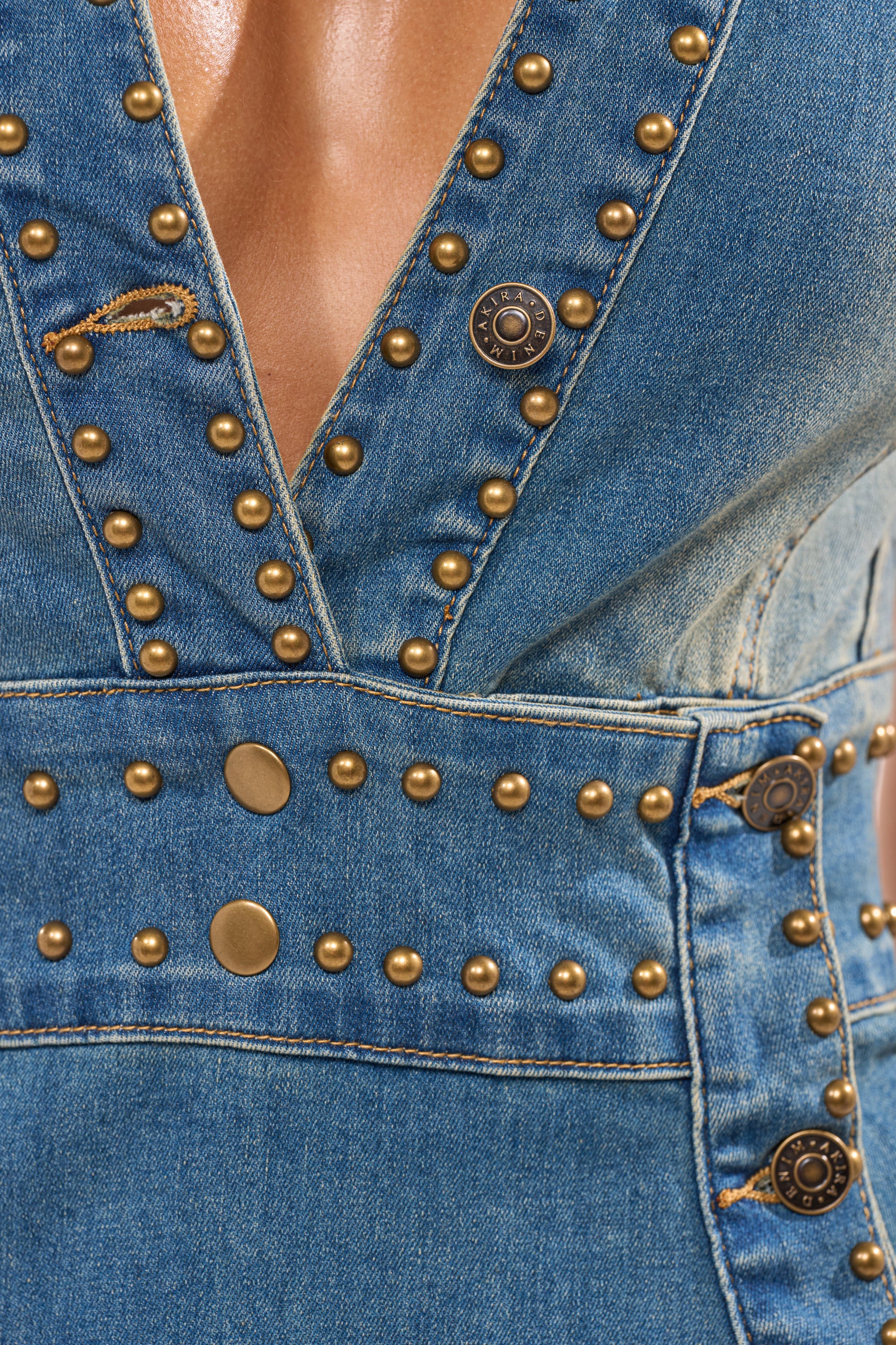 Close-up of the BABY GIRL STUDDED DENIM MINI DRESS, showcasing gold metal studs and buttons, a deep V-neckline, visible stitching, and a curve-hugging silhouette for a stylish touch on sturdy denim. 9