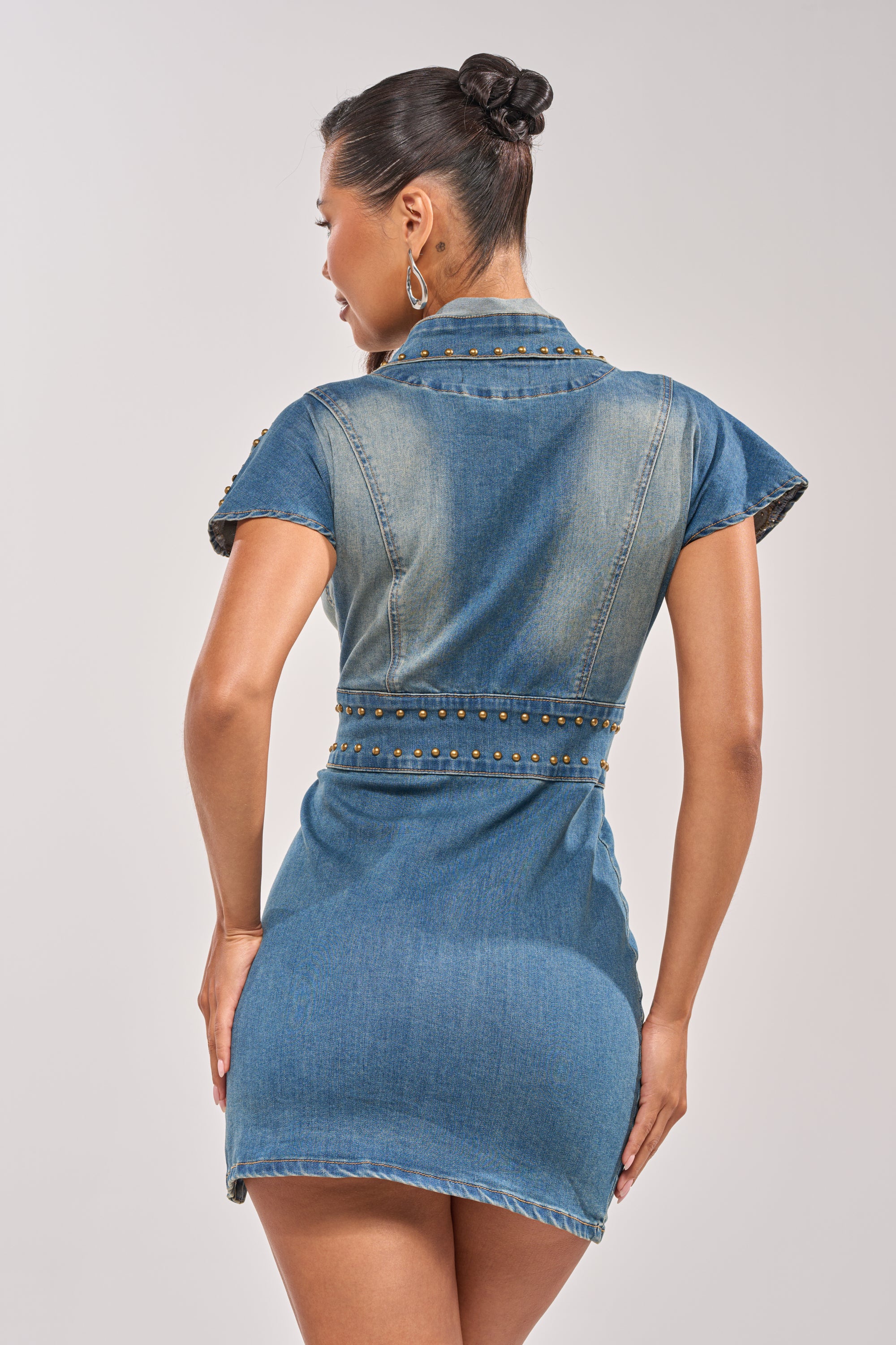A woman faces away, wearing the BABY GIRL STUDDED DENIM MINI DRESS—a curve-hugging, dark-wash mini dress with studded accents. Her hair is in a bun and she completes the look with large hoop earrings against a plain background. 6