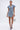 A woman poses against a plain background in the NEVER TOO MUCH DENIM MINI DRESS, featuring puffed sleeves, a front slit, and buttons. She completes her chic day-to-night look with hoop earrings, layered necklaces, a clear bag, and strappy blue heels.