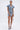 A woman stands against a plain background wearing the NEVER TOO MUCH DENIM MINI DRESS featuring short sleeves, an asymmetric hem, and button-up front, styled with strappy blue heels, a small purse, layered necklaces, and hoop earrings.