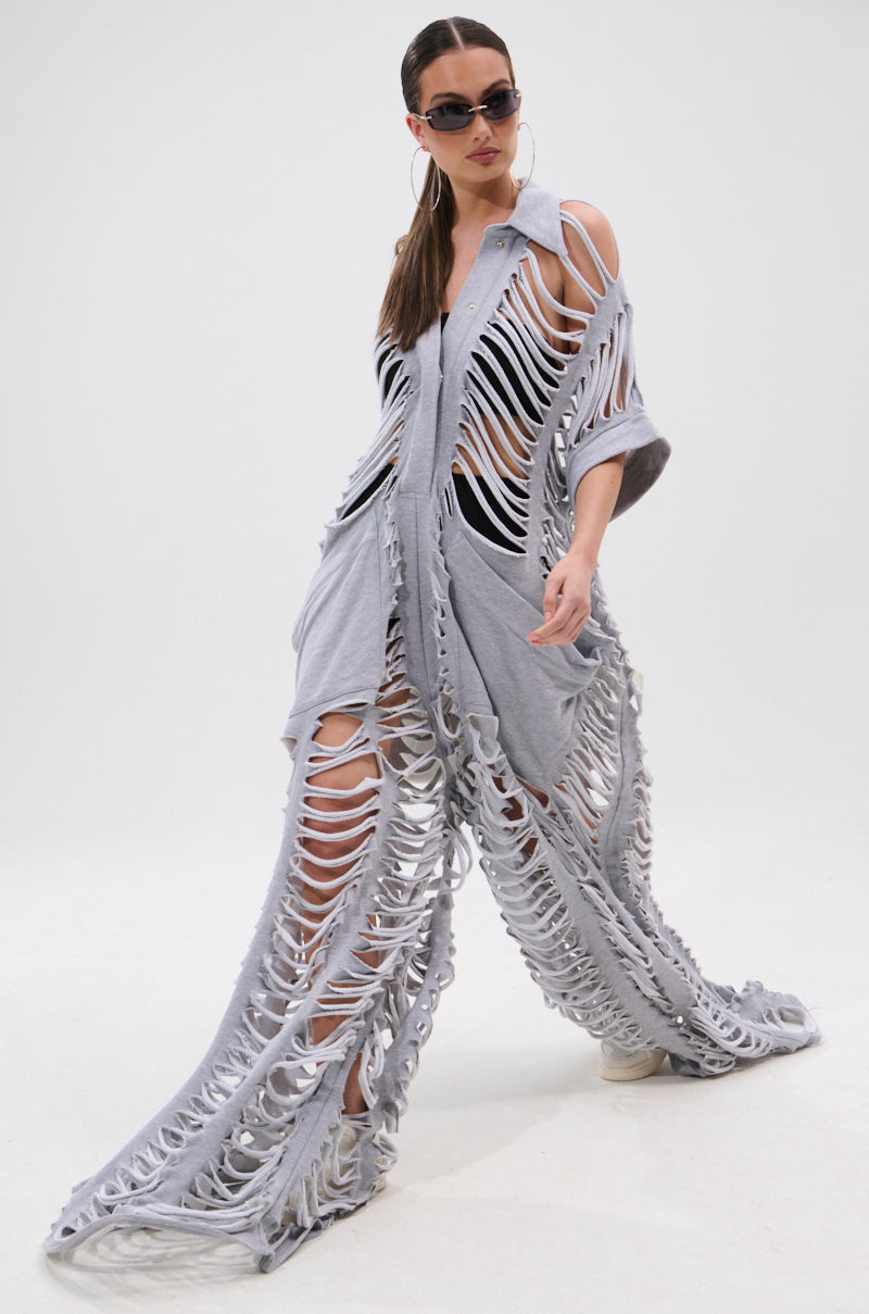A woman in sunglasses models the OVER IT DISTRESSED FRENCH TERRY JUMPSUIT in heather grey, featuring bold shredded cutouts, as she walks confidently against a plain white background.