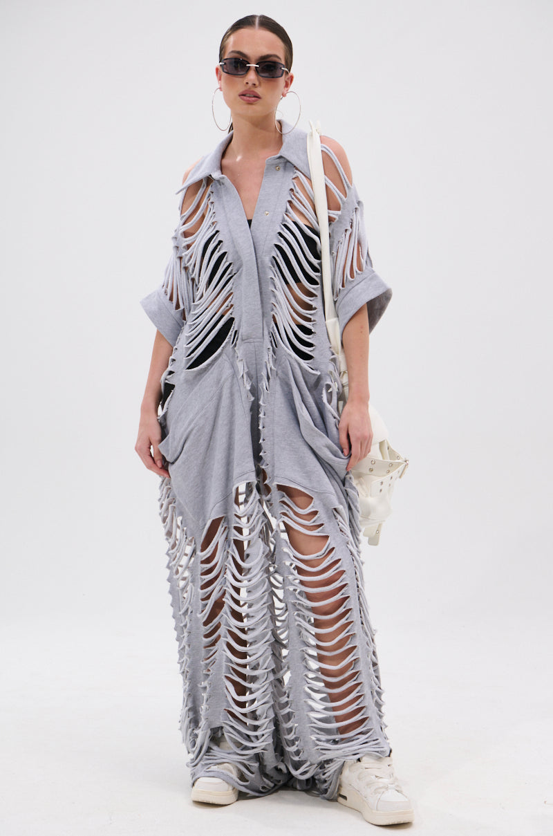 Woman wears the OVER IT DISTRESSED FRENCH TERRY JUMPSUIT in heather grey, featuring large shredded holes that reveal skin and black undergarments. She completes the look with sunglasses, hoop earrings, white sneakers, and a white bag.
