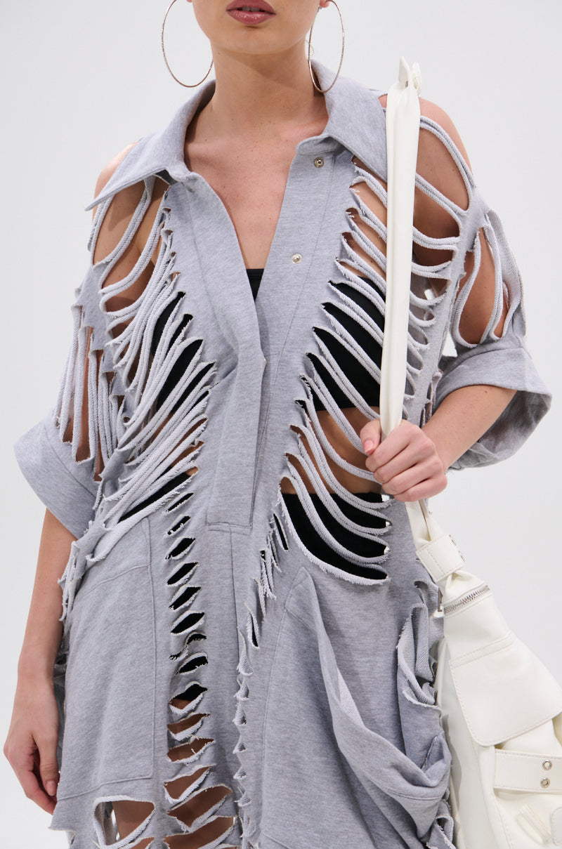 Model wears large hoop earrings and the OVER IT DISTRESSED FRENCH TERRY JUMPSUIT in heather grey with heavy cutouts, revealing a black undergarment, and holds a white bag against a plain white background.