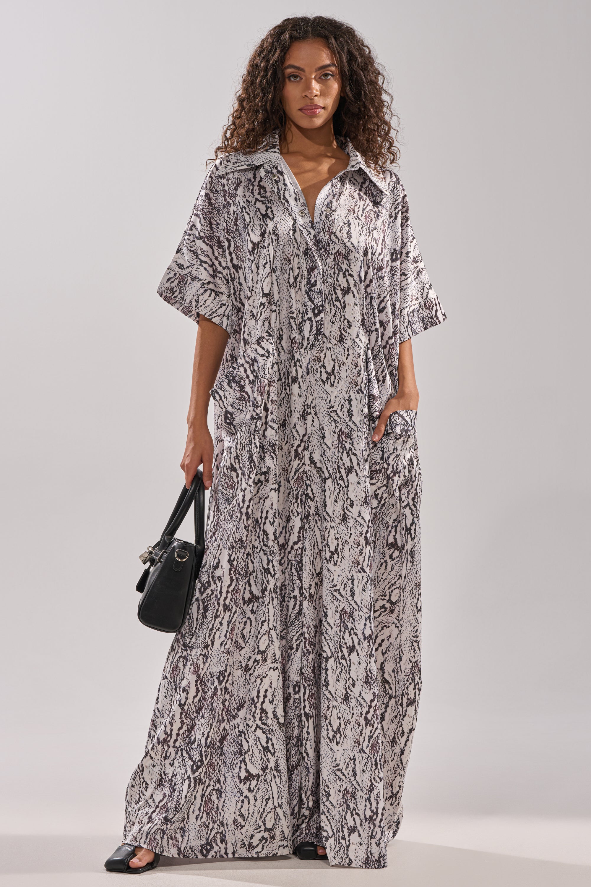 A woman with curly hair wears the SHE'S RICH Oversized Satin Jumpsuit in Snake Print, featuring short sleeves and a collar. She holds a black handbag and stands against a plain, light background.