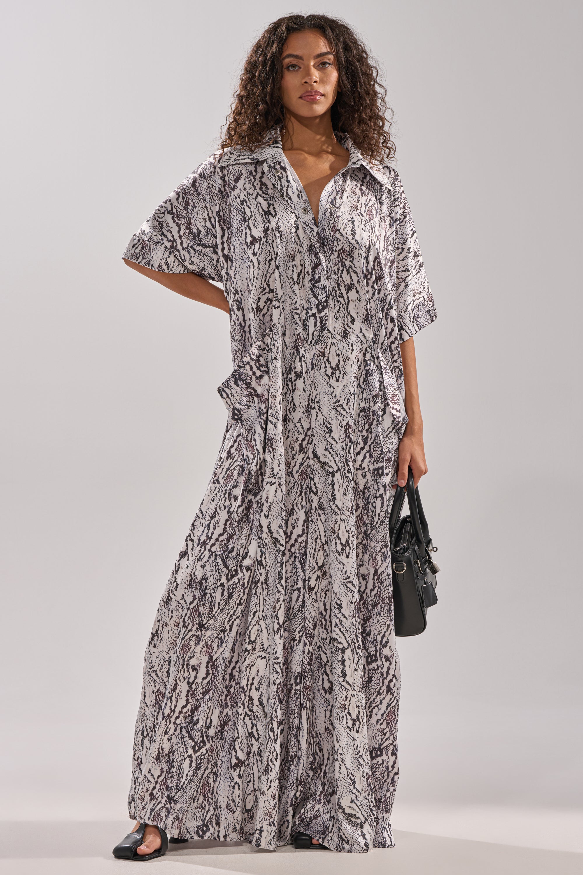 A woman with curly hair stands confidently in the SHE'S RICH OVERSIZED SATIN JUMPSUIT IN SNAKE PRINT, featuring a loose, floor-length fit. She accessorizes with a black handbag and black sandals against a plain, light background.