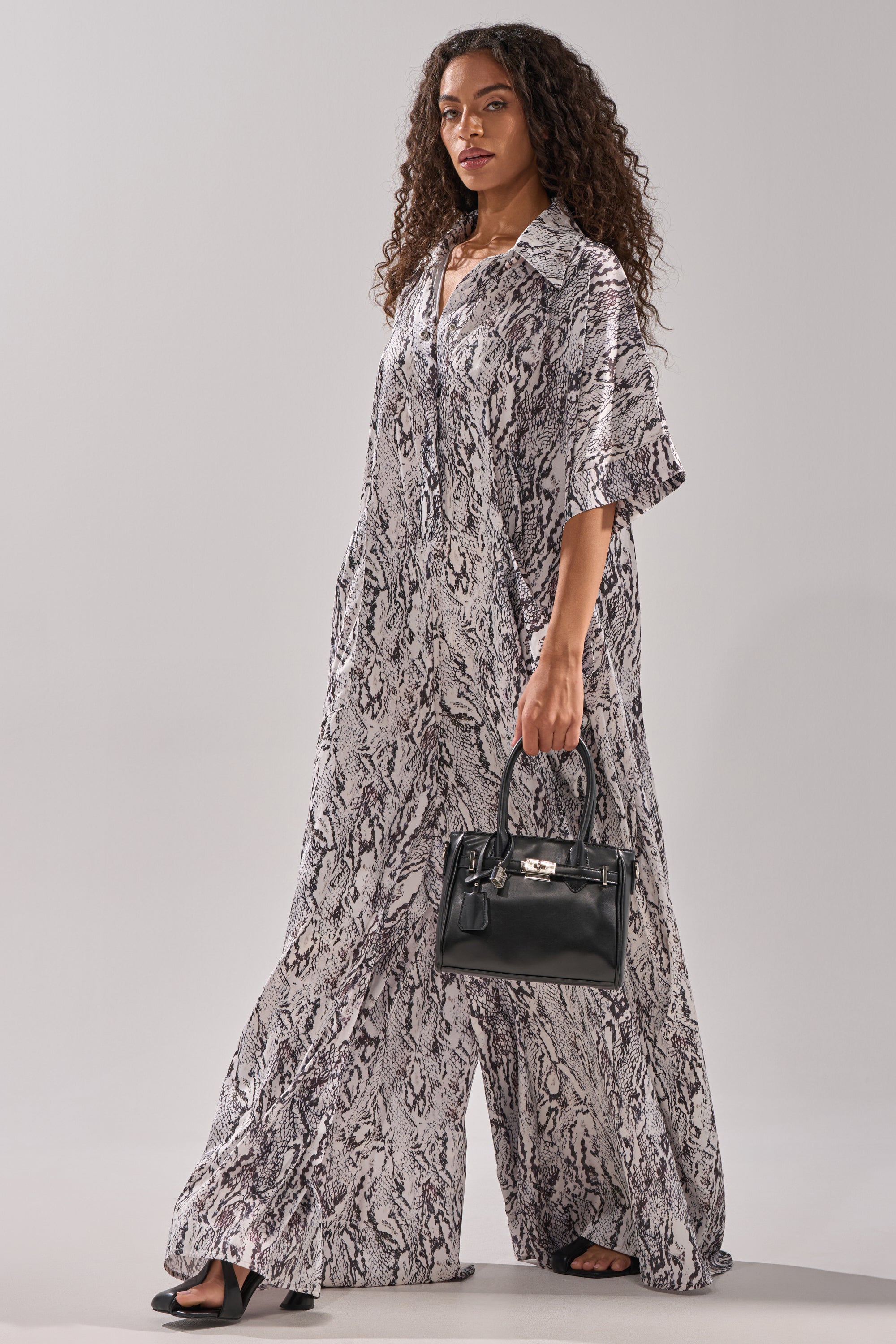 A woman with long curly hair poses against a light gray background, wearing the SHE'S RICH OVERSIZED SATIN JUMPSUIT IN SNAKE PRINT with black sandals and holding a small black handbag.