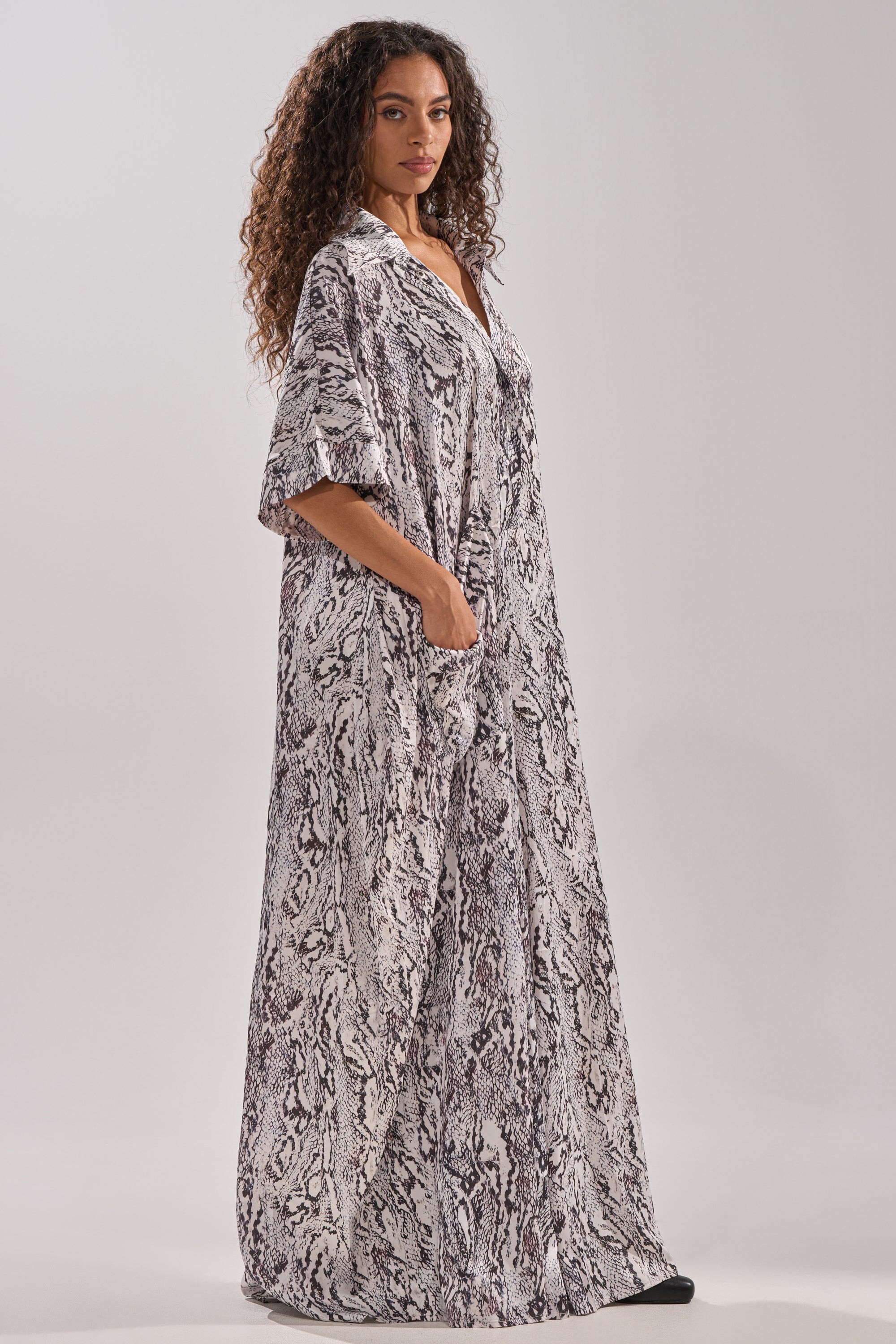 A woman with curly hair models the SHE'S RICH OVERSIZED SATIN JUMPSUIT IN SNAKE PRINT, featuring a loose fit, short sleeves, side pockets, and a black-and-white abstract pattern, as she poses against a plain background.