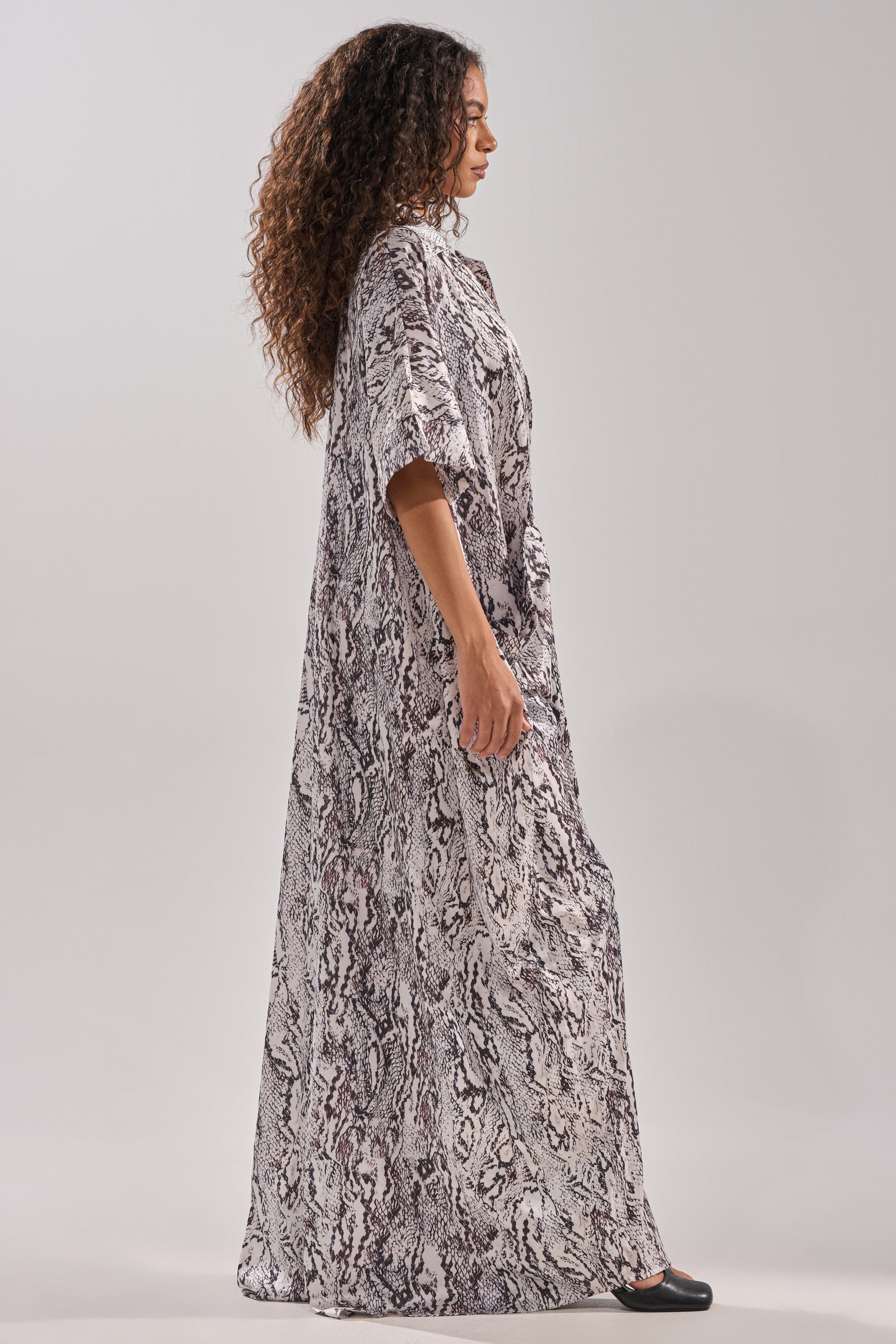 A woman with curly hair stands in profile against a light gray background, wearing the SHE'S RICH Oversized Satin Jumpsuit in snake print, featuring ankle-length gray and black tones, paired with black shoes.