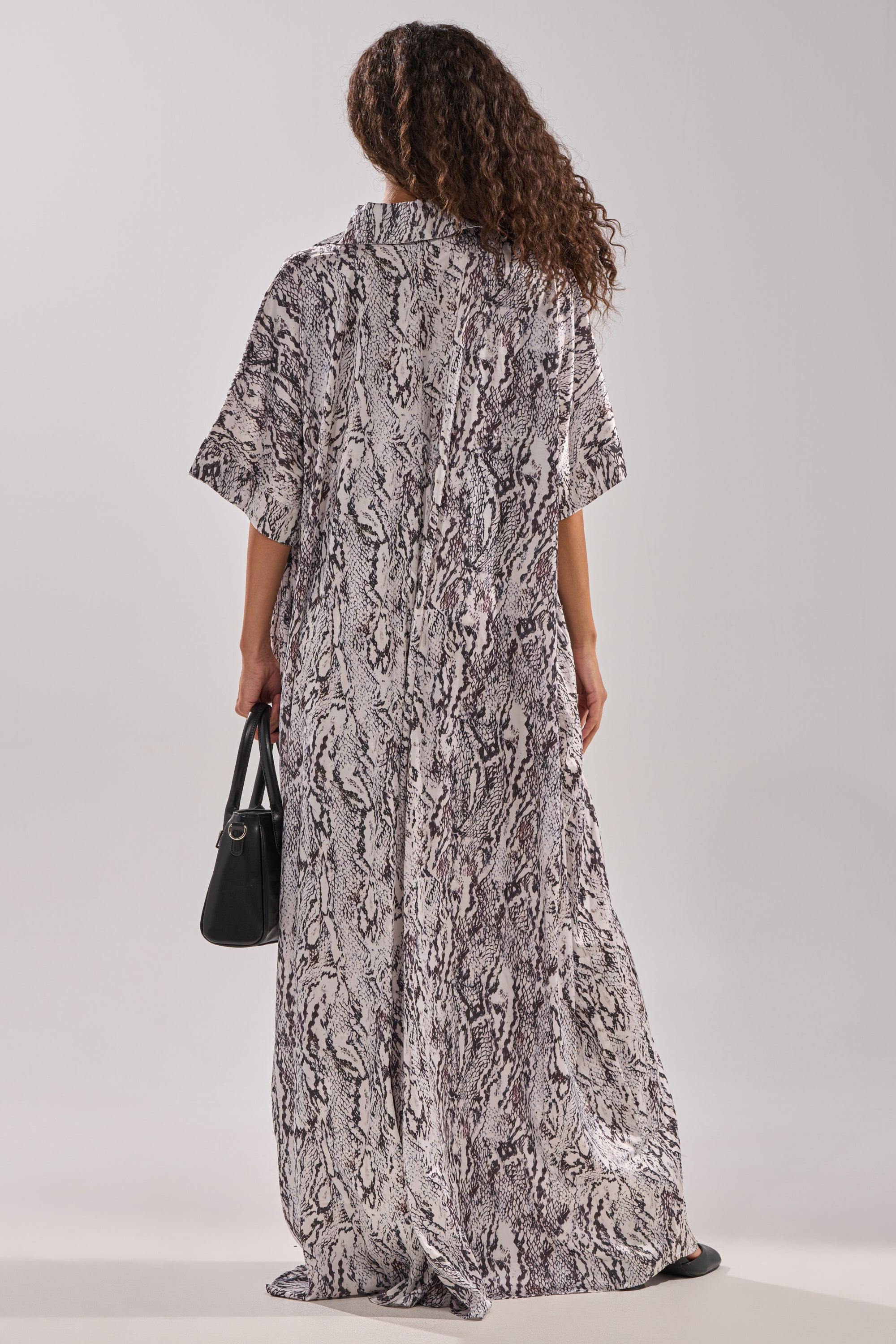 A person with curly hair, seen from behind, wears the SHE'S RICH OVERSIZED SATIN JUMPSUIT IN SNAKE PRINT with short sleeves and carries a black handbag against a plain light background.