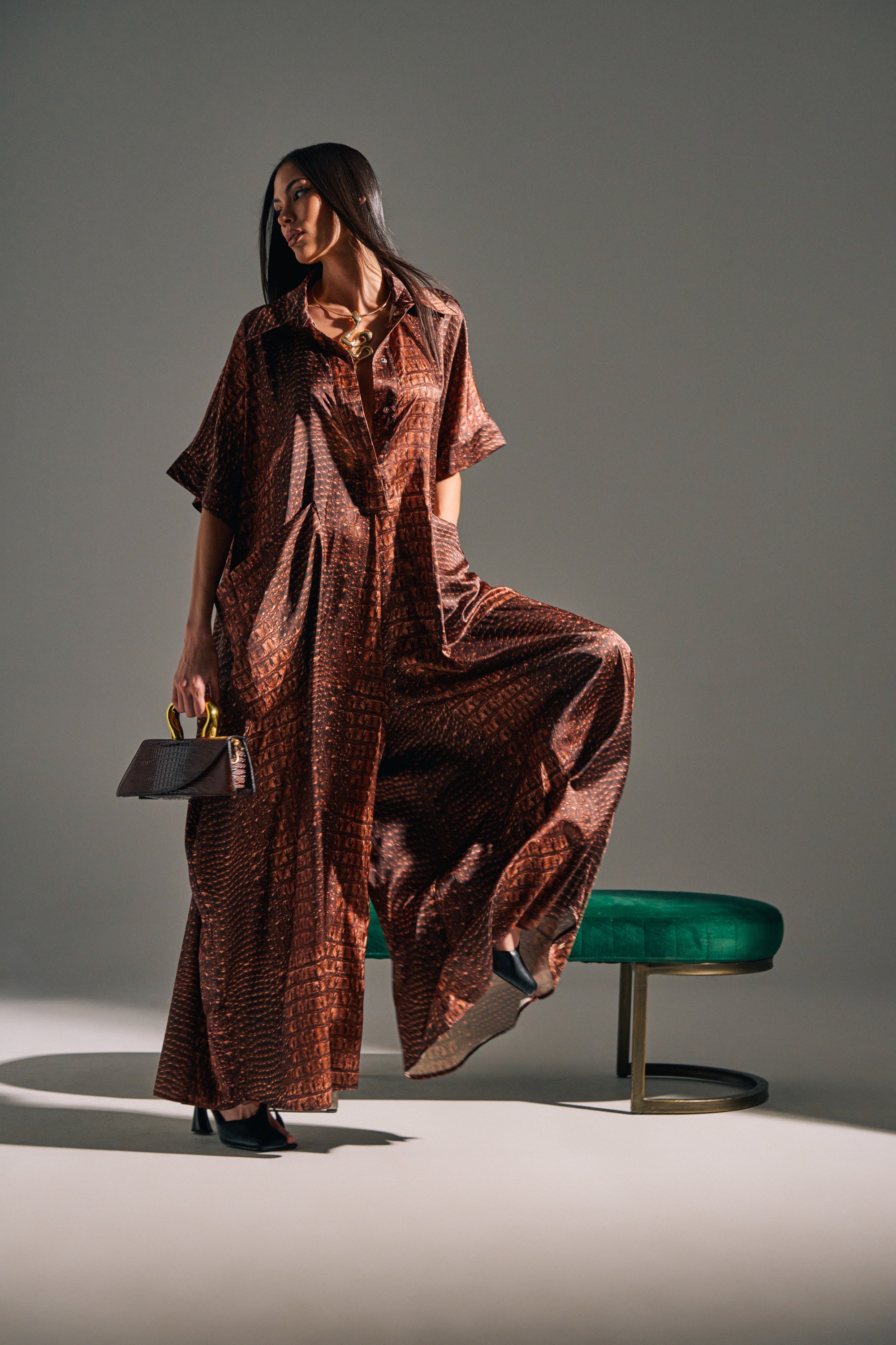 A woman poses confidently in the SHE'S RICH OVERSIZED SATIN JUMPSUIT IN CROC PRINT, holding a small handbag by a green bench under dramatic lighting that casts bold shadows on the floor.