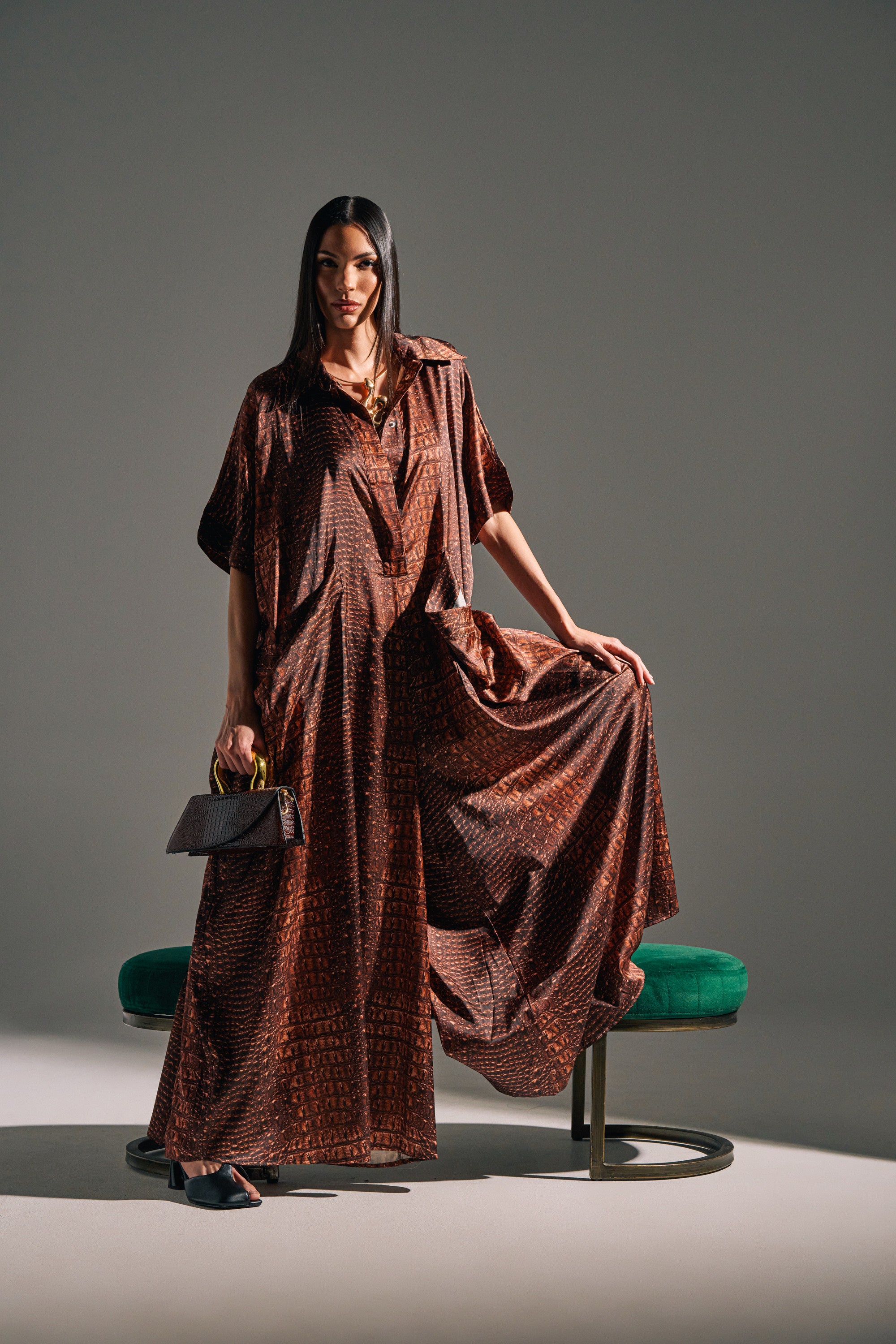 A woman poses confidently against a plain background, wearing the SHE'S RICH OVERSIZED SATIN JUMPSUIT IN CROC PRINT and holding a purse, with one foot on a green stool and part of her jumpsuit held up in one hand.