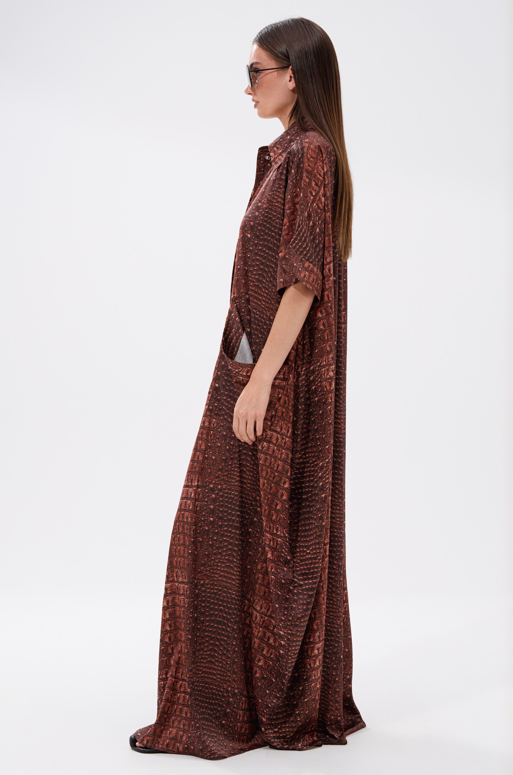 A woman in sunglasses and the AKIRA Label SHE'S RICH Oversized Satin Jumpsuit in Croc Print stands in profile against a plain white background.