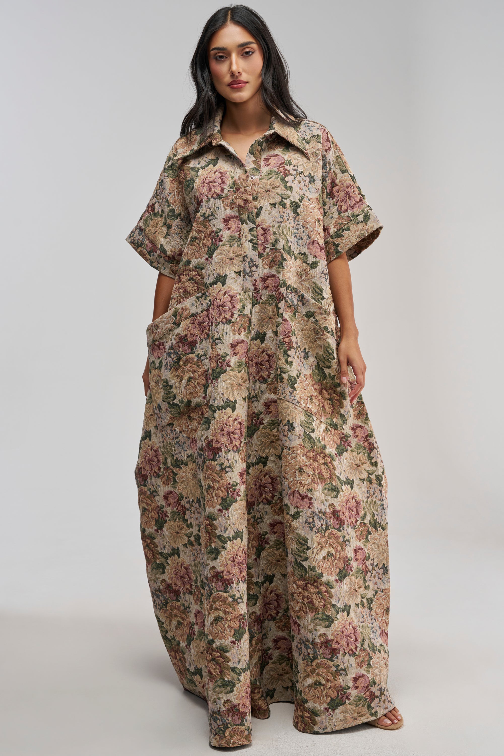 A woman stands wearing the VANJA BROCADE JUMPSUIT IN IVORY, a loose, floor-length style with short sleeves and a collar. Reminiscent of spring streetwear, this jumpsuit showcases a vintage-inspired floral pattern in shades of pink, green, and beige. She poses with her hands in the jumpsuit’s pockets.