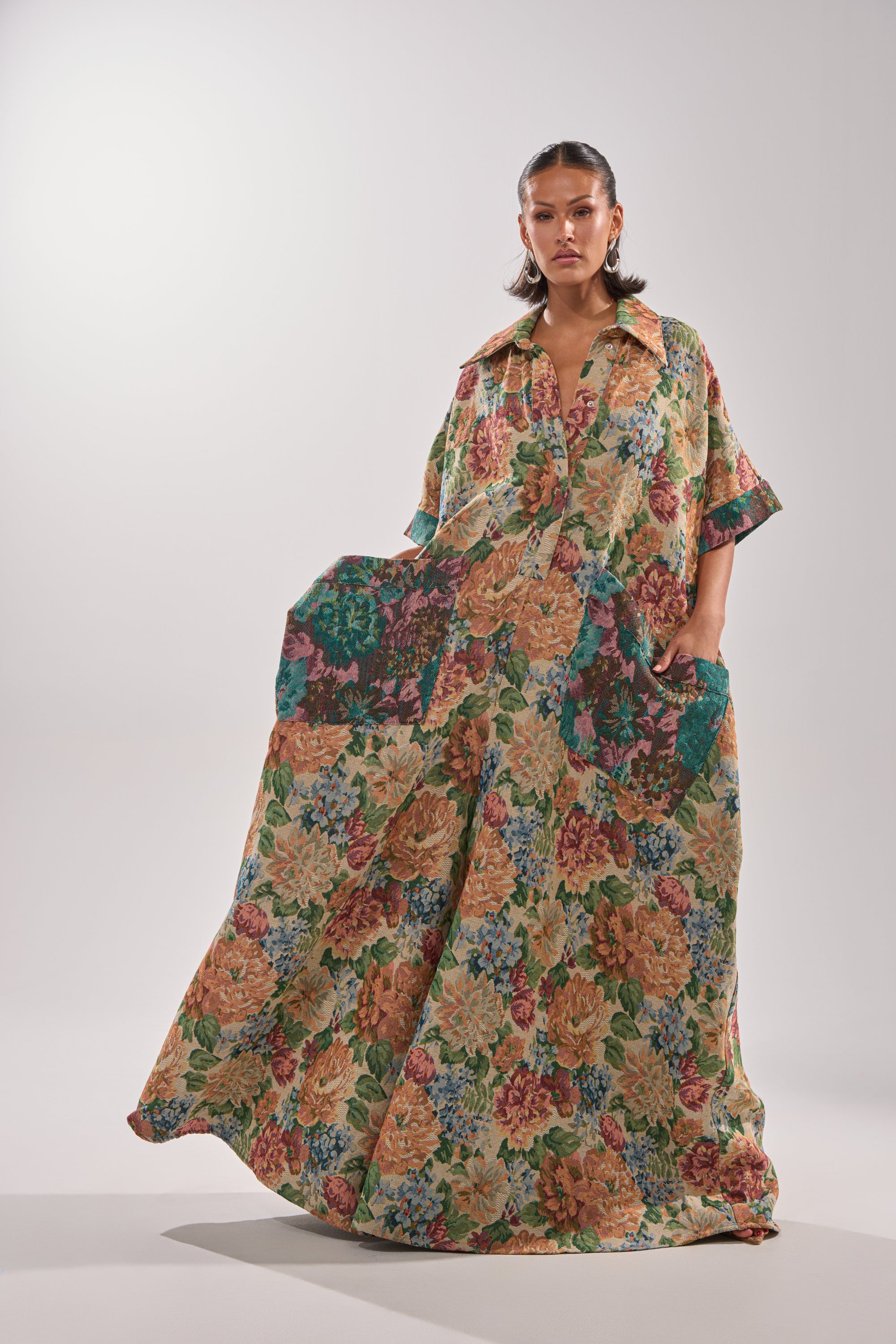 A woman stands confidently wearing the VANJA BROCADE JUMPSUIT, a long, loose-fitting one-piece featuring colorful floral tapestry-style fabric from shoulders to floor, set against a plain light background.
