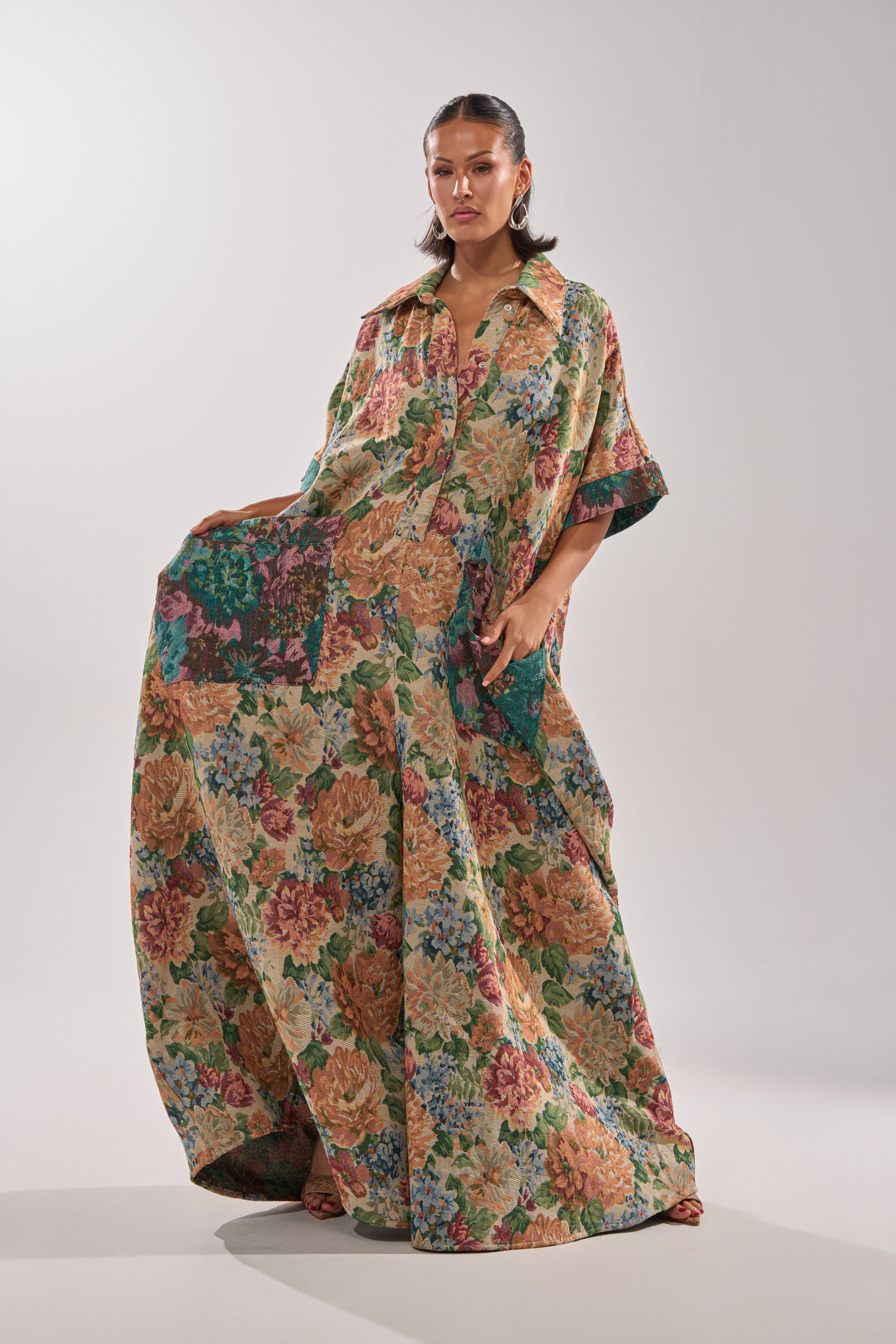 A woman stands confidently in the VANJA BROCADE JUMPSUIT, featuring wide sleeves, button front, large pockets, and a colorful oversized floral print in tapestry-style fabric. She poses with one hand in a pocket against a plain background.